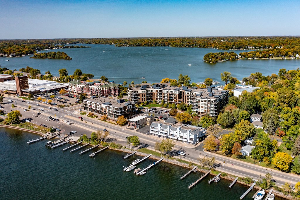 Aerial view of a lakeside town near The Mist on Lake Minnetonka Apartment Homes with docks, boats, and waterfront buildings. Surrounded by lush green trees and a calm blue lake under a clear sky.