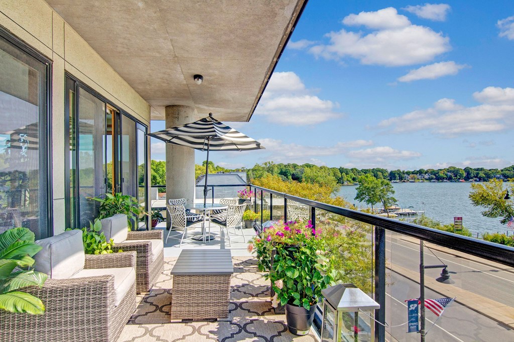 Modern balcony here at The Mist on Lake Minnetonka Apartment Homes overlooking a lake, featuring wicker furniture, a striped umbrella, and potted plants. The scene is bright and tranquil, evoking relaxation.