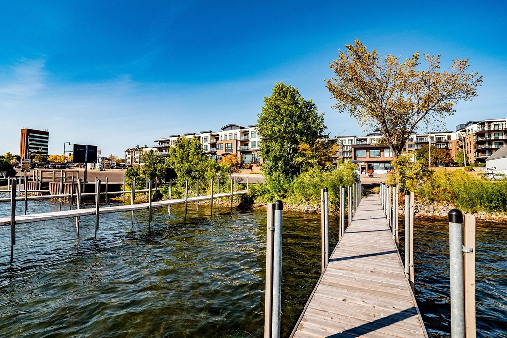 Wooden pier near The Mist on Lake Minnetonka Apartment Homes leading to a modern waterfront neighborhood with greenery and trees. Clear blue sky, calm water, and urban buildings convey tranquility.