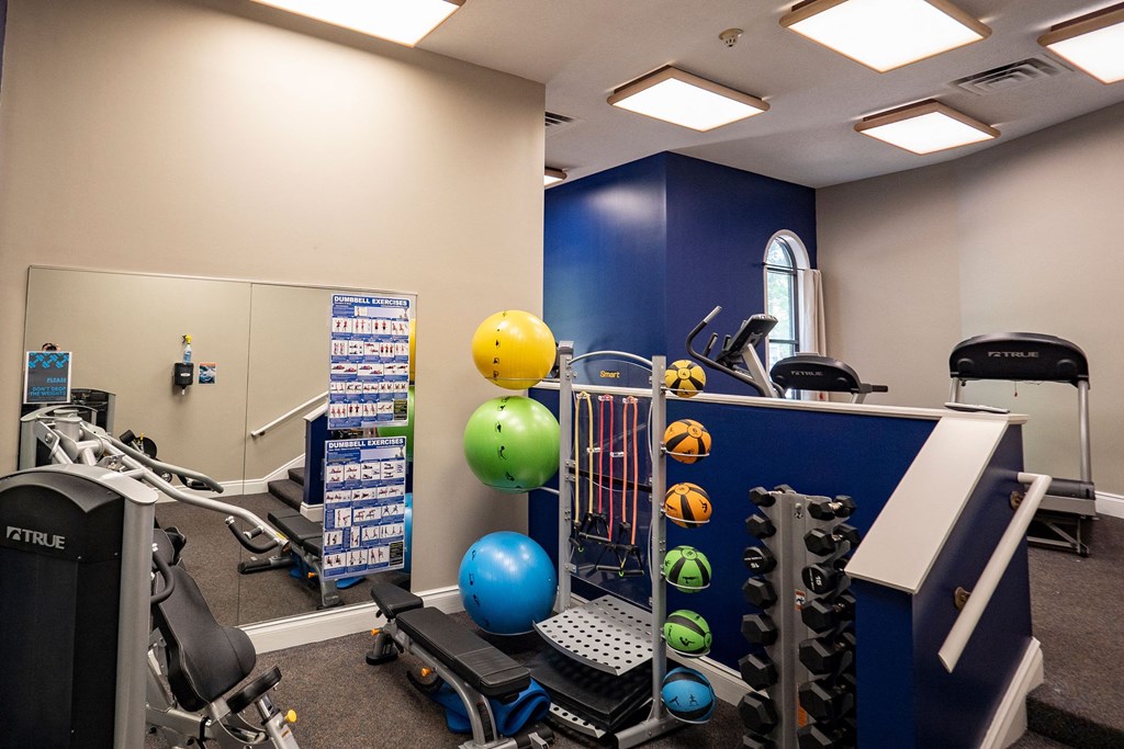 Plymouth Square Fitness Center A compact gym room here at Plymouth Square Apartment Homes with exercise equipment, colorful stability balls, a dumbbell rack, and workout posters. Mirrors and blue accents create a focused atmosphere.