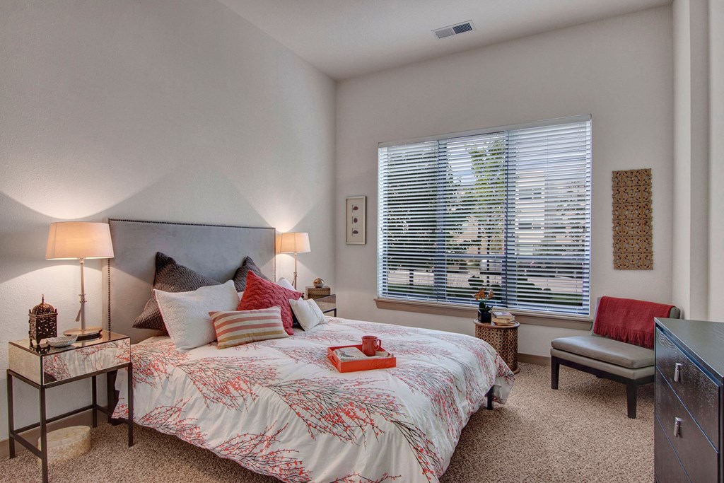 Bright bedroom here at The Residence at The COR Apartment Homes with soft lighting. Bed with floral duvet, assorted pillows, and bedside tables. Large window with blinds. Calm and inviting atmosphere.