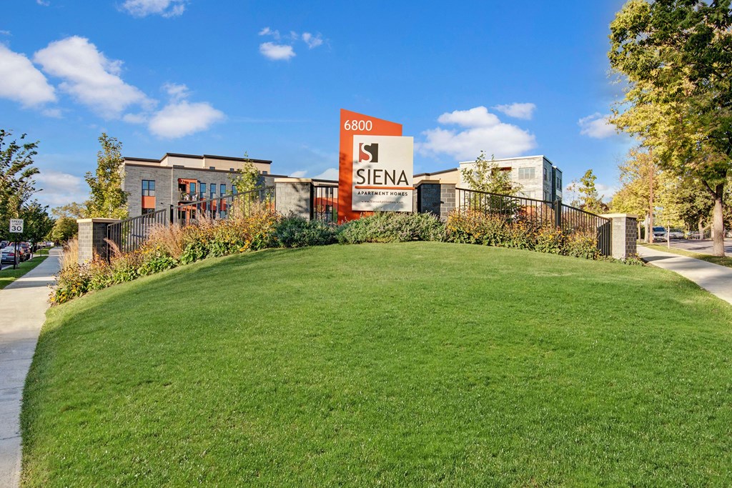 Siena Exterior A grassy hill leads to the Siena Apartment Homes entrance sign, surrounded by lush landscaping. Modern building and clear blue sky in the background.
