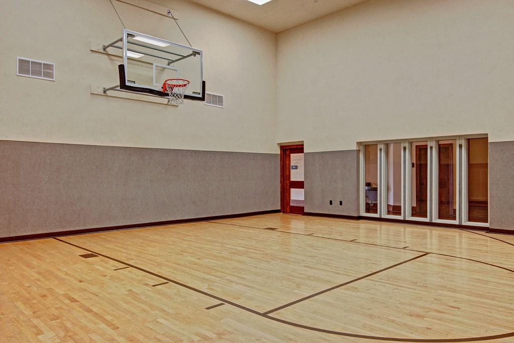 Indoor basketball court here at Stoneleigh at The Reserve Apartment Homes with a wooden floor, gray walls, and a basketball hoop mounted on the wall. Nearby is a closed door and glass panels.
