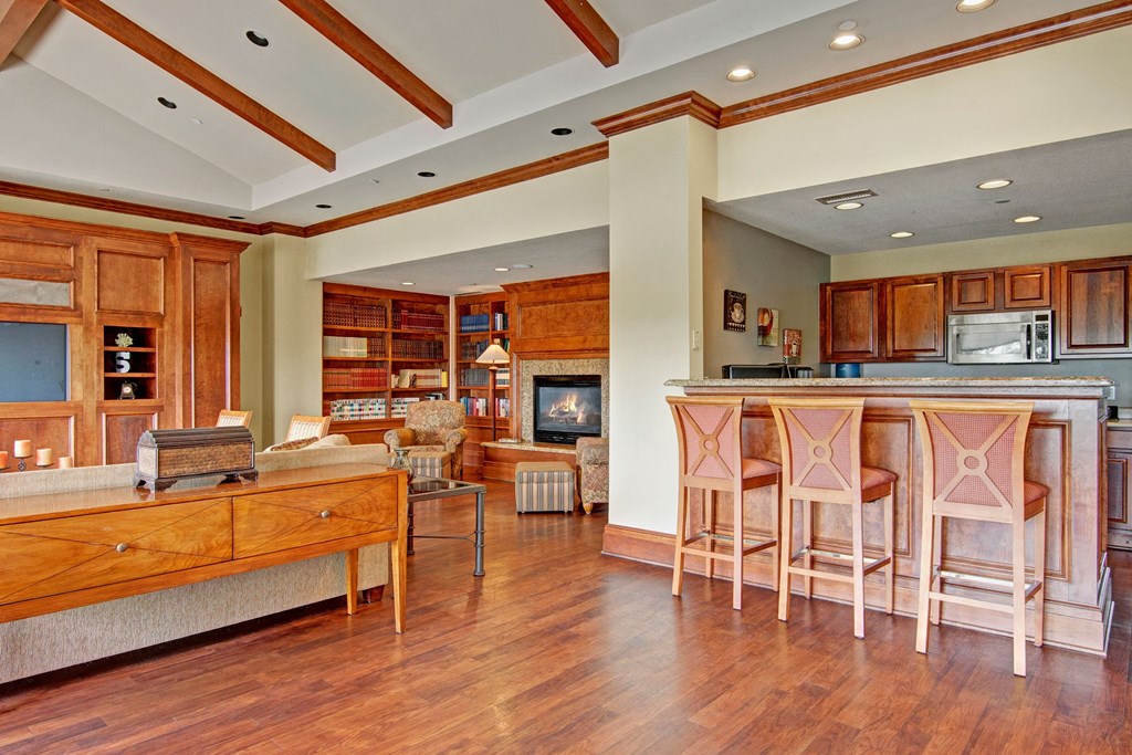 Warm, cozy resident lounge here at Stoneleigh at The Reserve Apartment Homes with wooden furniture, a brick fireplace, and bookshelves in the background. The kitchen has a sleek island and bar stools.
