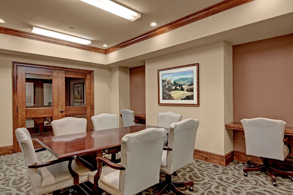 Elegant conference room here at Stoneleigh at The Reserve Apartment Homes with a polished wood table, plush white chairs, beige walls, landscape painting, and soft lighting, conveying a professional atmosphere.