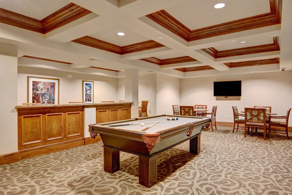 Luxurious recreation room here at Stoneleigh at The Reserve Apartment Homes with a pool table, ornate carpet, dark wood paneling, and ceiling beams. Art on walls, TV, and dining sets create a cozy, elegant vibe.