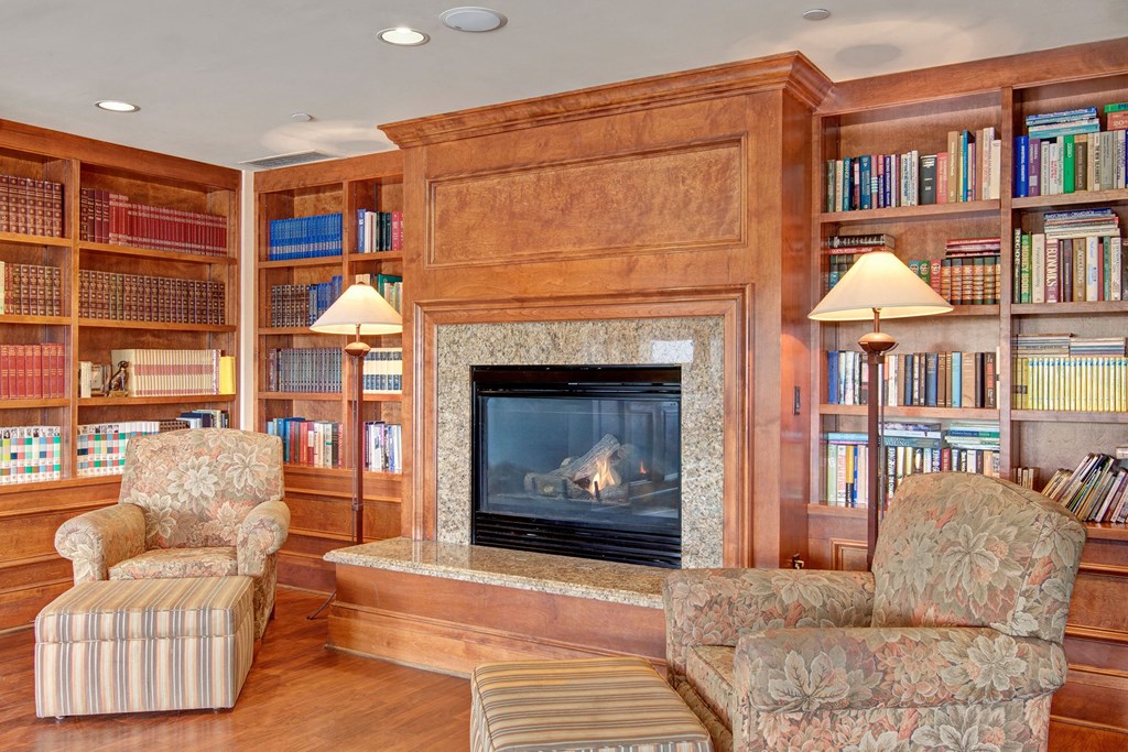 Cozy library here at Stoneleigh at The Reserve Apartment Homes with wooden shelves filled with colorful books, a lit fireplace, and two patterned armchairs with a footrest, creating a warm atmosphere.
