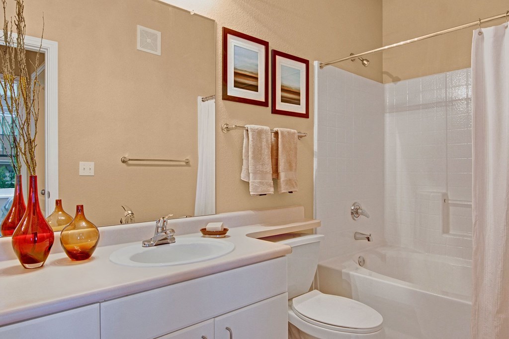 Stylish bathroom here at Stoneleigh at The Reserve Apartment Homes featuring a white vanity with a round sink, red and amber vases, beige walls, framed artwork, and a bathtub with a shower curtain.