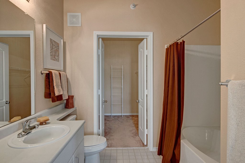 Bathroom here at Stoneleigh at The Reserve Apartment Homes with beige walls, a white sink with soap, and a toilet. Maroon shower curtain and towels. Door leads to a carpeted closet with white shelves.