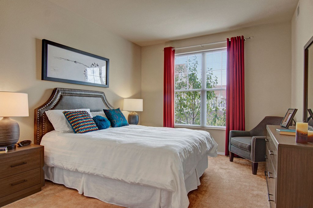 A cozy bedroom here at Stoneleigh at The Reserve Apartment Homes with a large bed featuring a studded headboard and colorful cushions. Two lamps on nightstands and a window with red curtains enhance the warm ambiance.