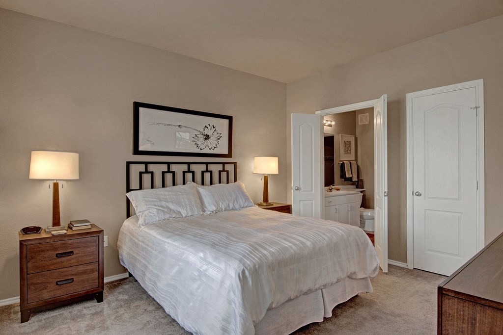 A cozy bedroom here at Stoneleigh at The Reserve Apartment Homes with a neatly made double bed, flanked by two wooden nightstands with lamps. A simplistic floral artwork adorns the beige wall.