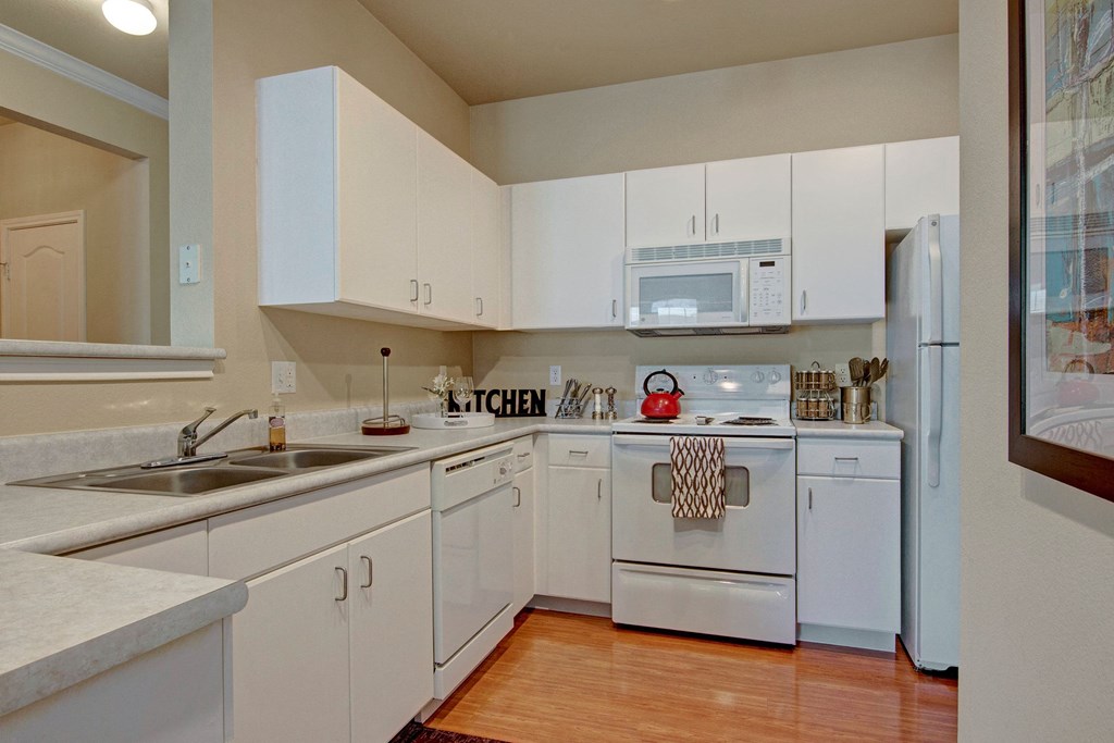 A bright kitchen here at Stoneleigh at The Reserve Apartment Homes with white cabinets, countertops, and appliances. A red kettle on the stove adds a pop of color. The atmosphere is clean and modern.