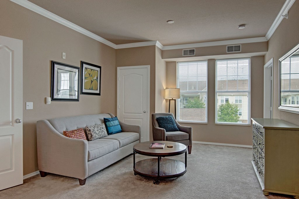 Cozy living room here at Stoneleigh at The Reserve Apartment Homes with beige walls, a gray sofa, and a blue accent chair. Large windows let in natural light. Two framed artworks hang above the sofa.