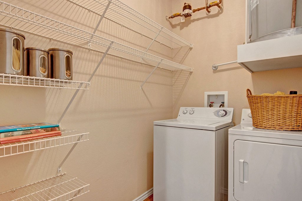 StoneleiA small laundry room here at Stoneleigh at The Reserve Apartment Homes with a white washer and dryer on the right. Wire shelving on the left holds metal canisters and cookbooks. A wicker basket sits atop the dryer.gh Spacious Laundry Area