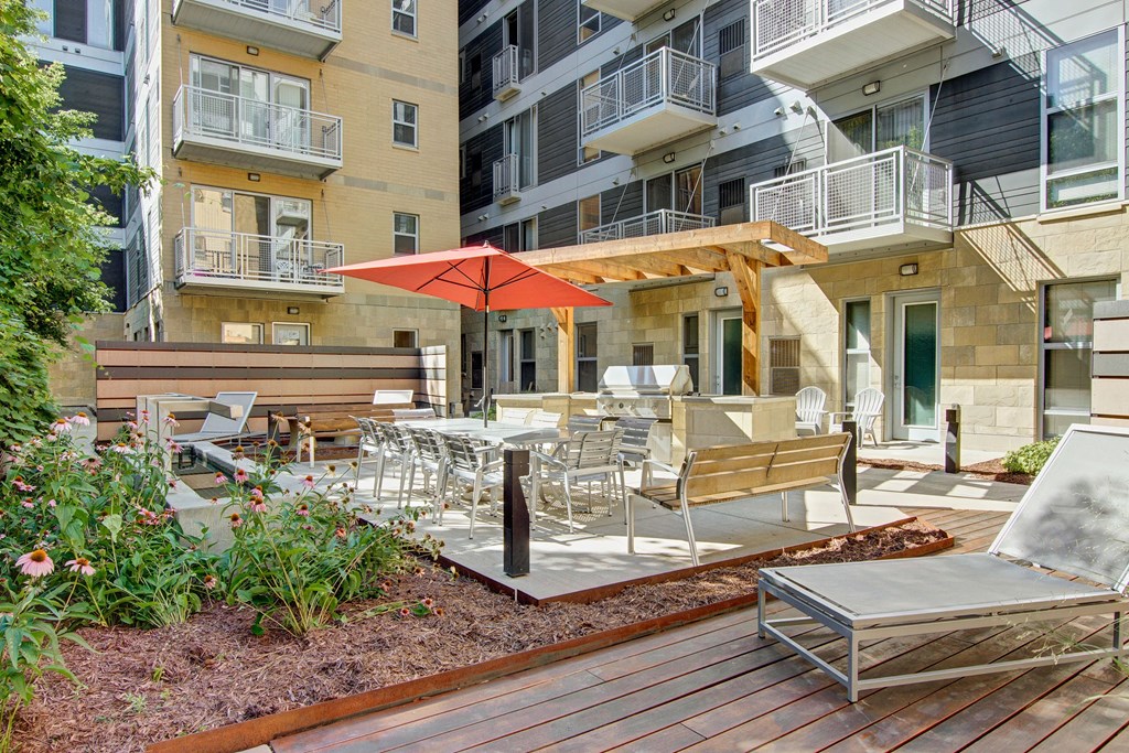 Modern courtyard here at VUE Apartment Homes with seating area, dining set under a red umbrella, grill, lounge chairs, and flower beds. Bright and inviting ambiance.