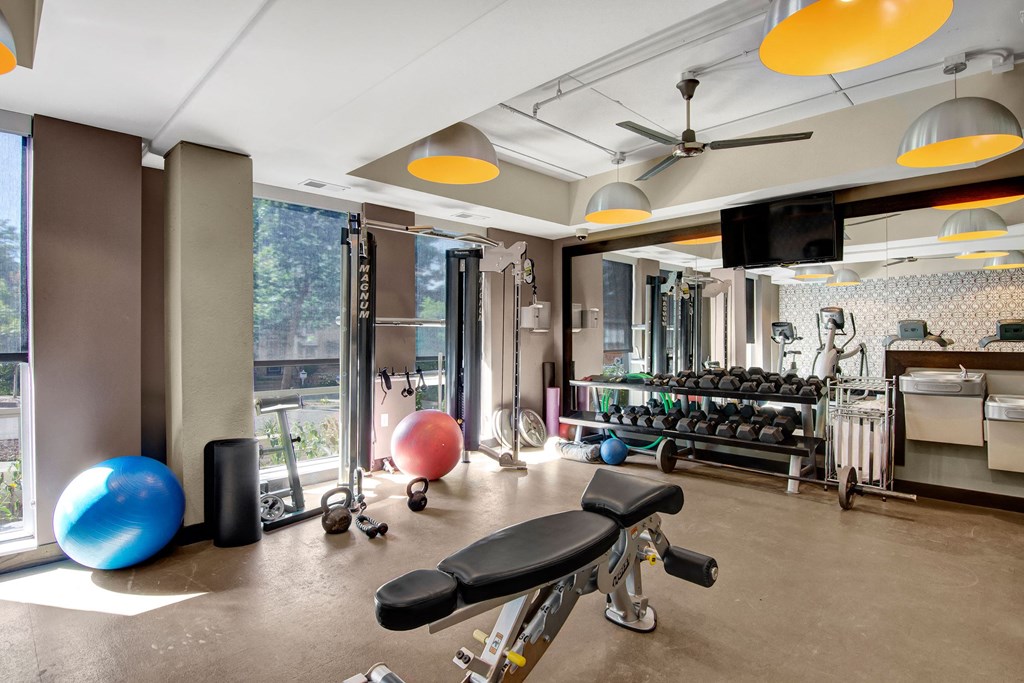 Bright gym here at VUE Apartment Homes with large windows, exercise machines, dumbbells, and yoga balls. Modern decor with hanging lights creates an energetic, welcoming atmosphere.