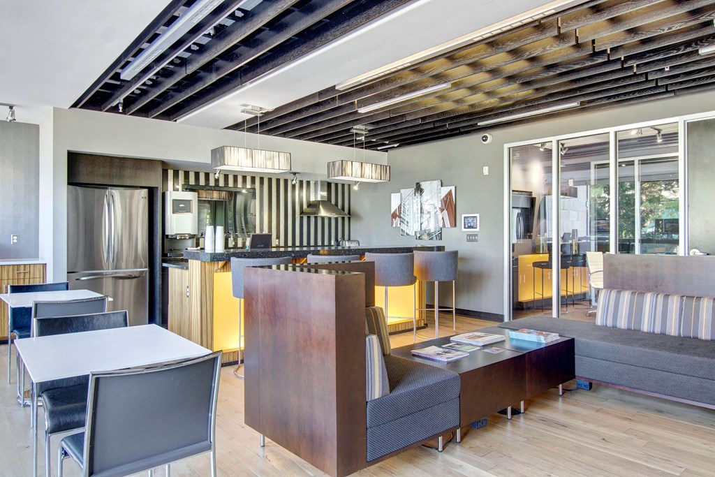 Modern kitchen and lounge area here at VUE Apartment Homes with sleek design. Features stainless steel fridge, bar with chairs, pendant lights, striped sofa, and wood accents. Cozy and stylish atmosphere.