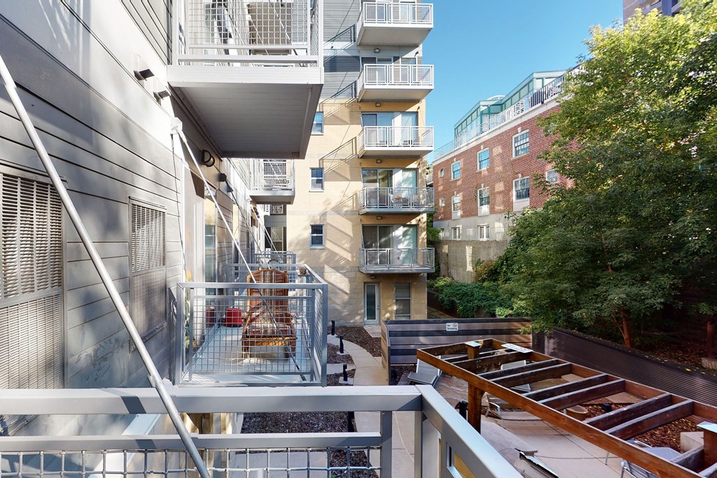 Elevated view of our modern VUE Apartment Homes complex. Metal balconies line the facade, adjacent to a red brick building. Trees and a clear blue sky in the background.
