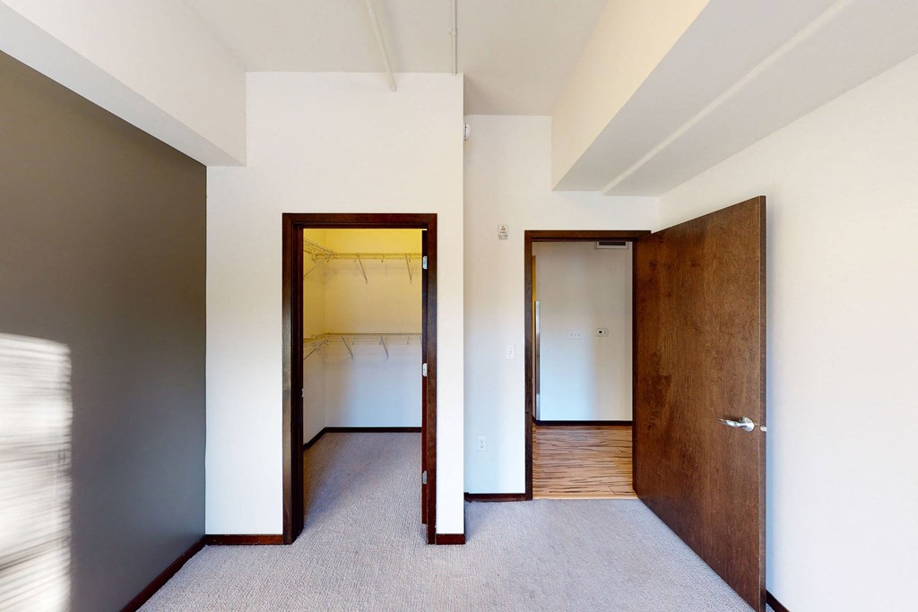 Empty room here at VUE Apartment Homes with beige walls and carpeted floor, featuring two open wooden doors; one leads to a walk-in closet, the other to a hallway with wood flooring. Bright and minimalist.
