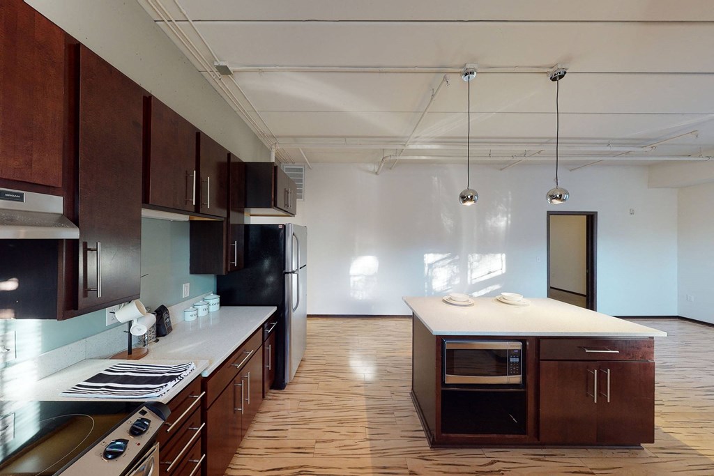 Spacious kitchen here at VUE Apartment Homes with dark wood cabinets, stainless steel appliances, and a central island with hanging lights. Natural light streams through windows.