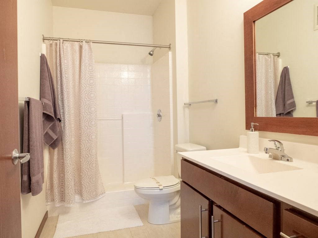 Modern bathroom here at VUE Apartment Homes with a wooden vanity and white countertop. A toilet and shower with a patterned curtain are in the background. Neutral tones create a calm ambiance.