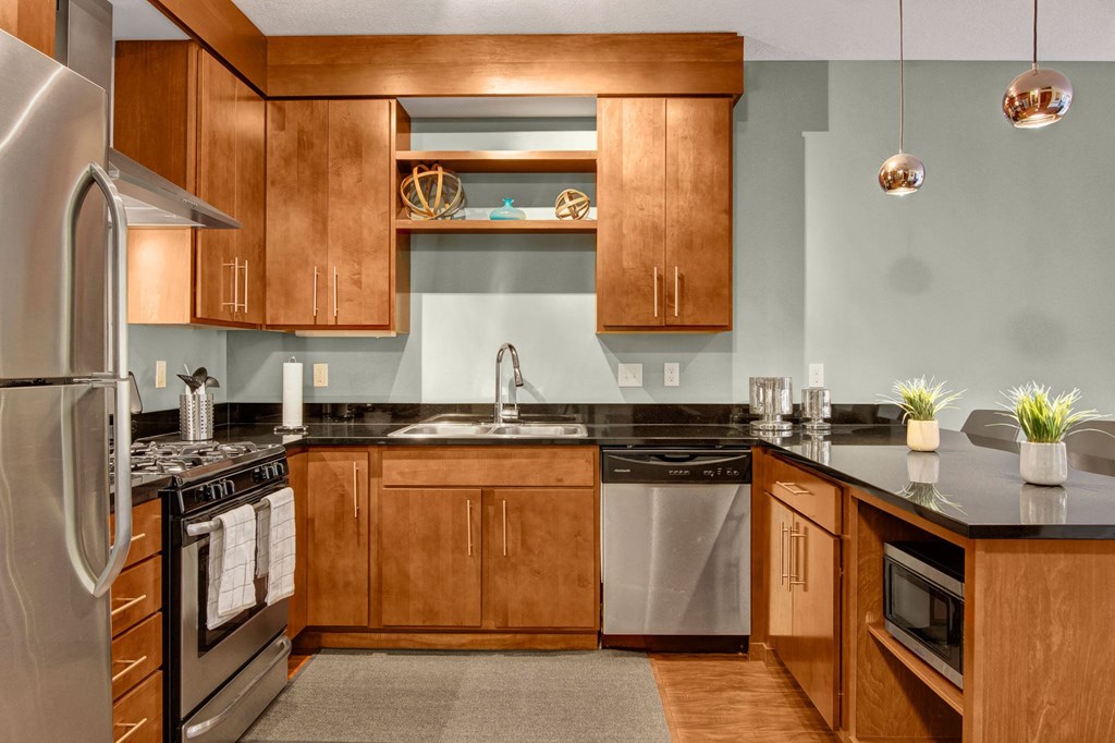 Modern kitchen here at VUE Apartment Homes with wooden cabinets, stainless steel appliances, and black countertops. Hanging pendant lights and potted plants add a cozy feel.