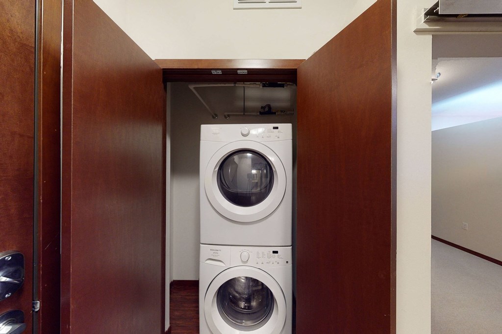 Stacked white washer and dryer in a small closet here at VUE Apartment Homes with wooden doors, conveying a tidy and compact laundry area. Brown floor and beige walls visible.