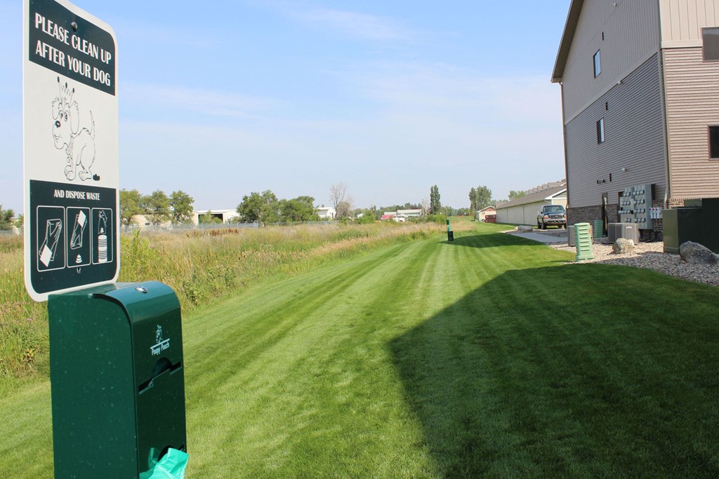 Sign urging dog owners to clean up waste is posted on a grassy path bordered by a building and a field here at Dakota Commons Apartment Homes. The sunny day conveys a pleasant atmosphere.