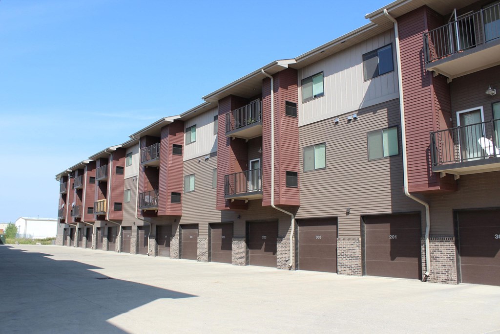 Three-story Dakota Commons Apartment Homes complex with brown siding and brick accents. Each unit has a balcony and a garage. Clear blue sky suggests a sunny day.