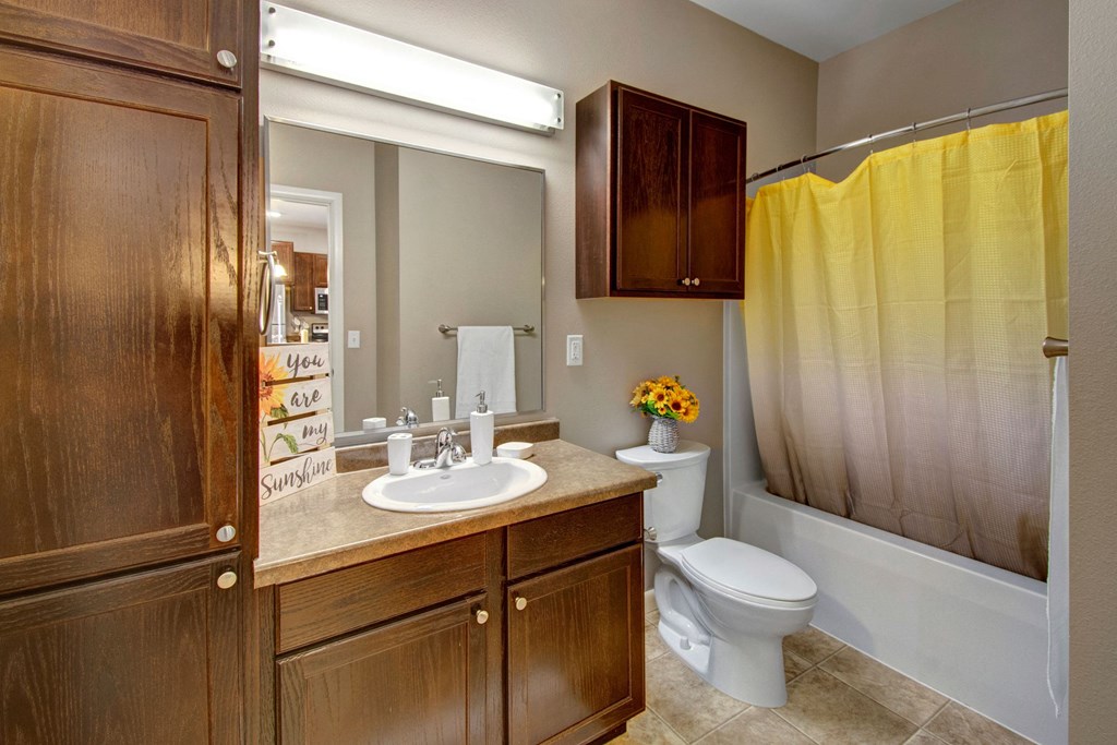 A cozy bathroom here at Dakota Commons Apartment Homes with wooden cabinets, a sink, mirror, and beige countertop. A bright yellow shower curtain adds warmth beside a toilet with a sunflower bouquet.