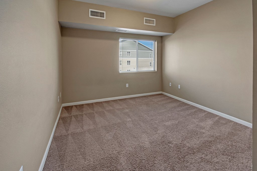 Empty bedroom here at Dakota Commons Apartment Homes with beige walls and carpet, featuring a window with blinds. Soft natural light enters, creating a calm, neutral atmosphere.