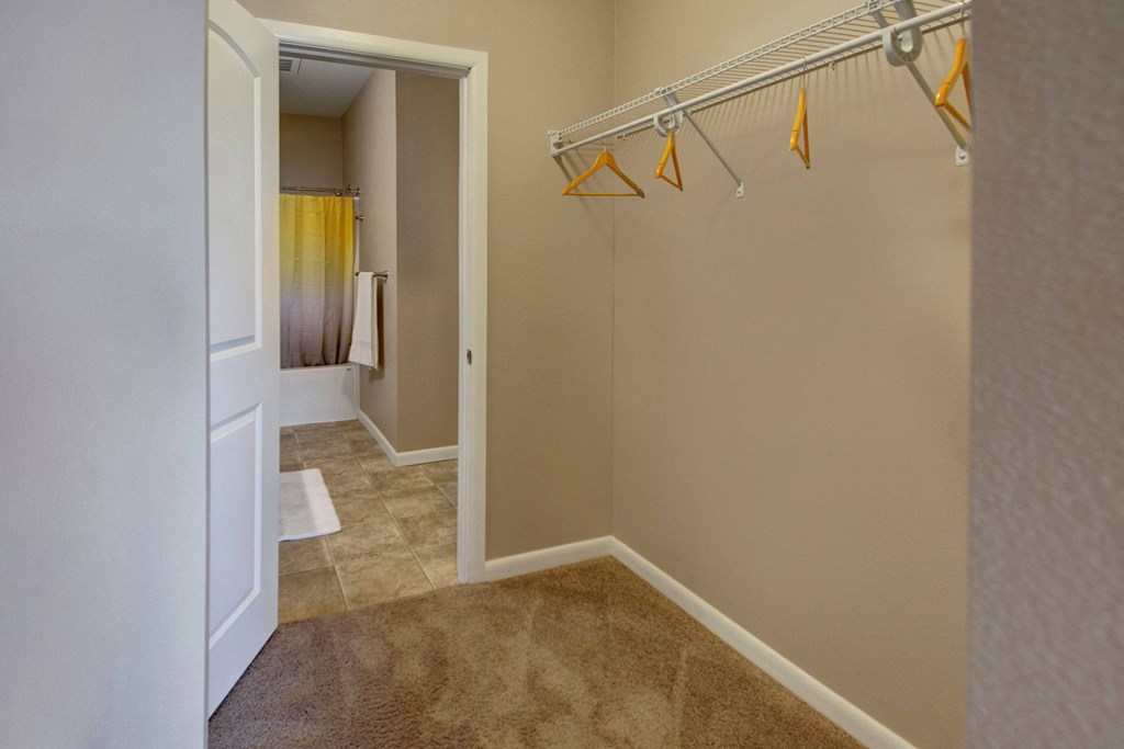 Walk-in closet here at Dakota Commons Apartment Homes with beige walls and brown carpet, featuring empty wooden hangers on a wire rack. An open door reveals a bathroom with a yellow gradient curtain.