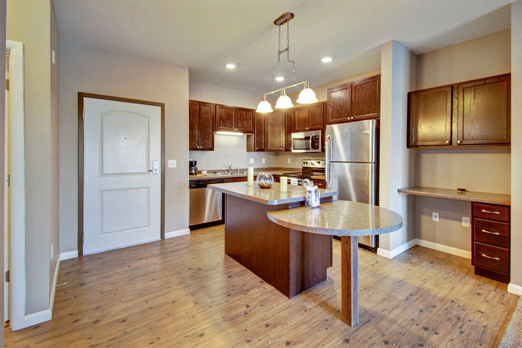 Modern kitchen here at Dakota Commons Apartment Homes, with wooden floors, a central island, stainless steel appliances, and dark wood cabinets. Bright lighting creates a warm, inviting feel.