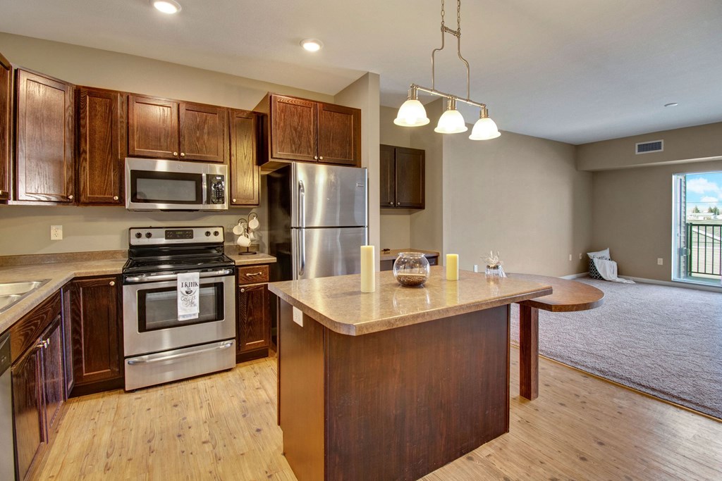 Modern kitchen here at Dakota Commons Apartment Homes, with dark wood cabinets, stainless steel appliances, and an island with pendant lighting. Open living area with carpet and balcony view.