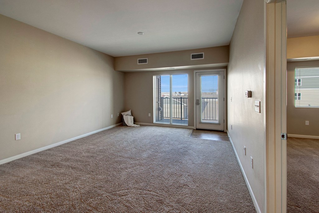 A bright, unfurnished living room here at Dakota Commons Apartment Homes features beige walls and carpet. Glass doors open to a small balcony. A single pillow rests in the corner, creating a serene, open atmosphere.