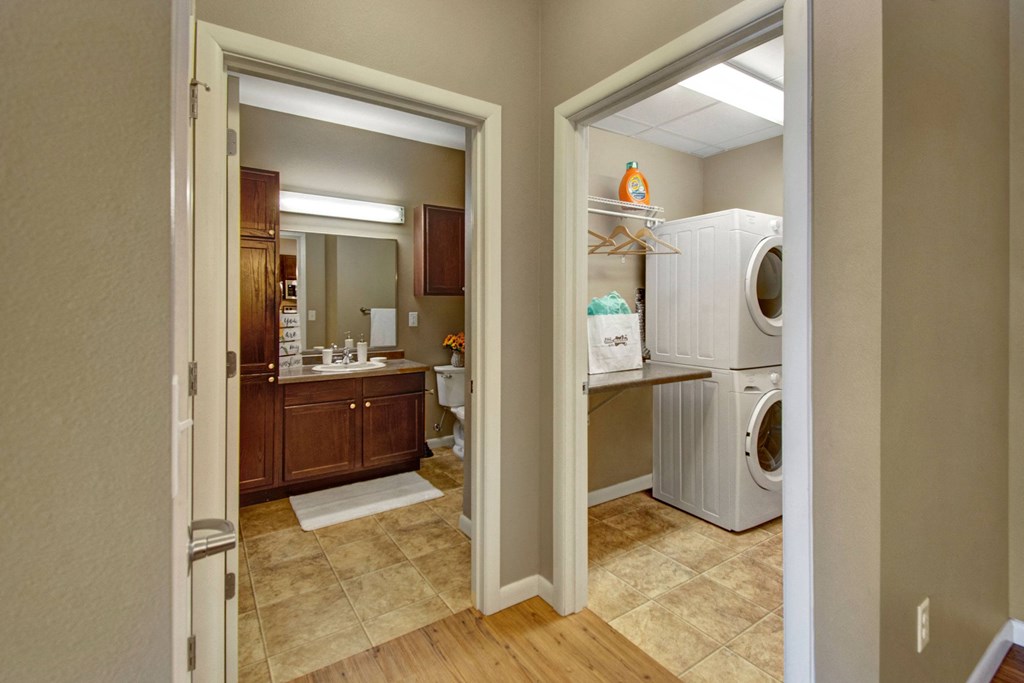 A warm-toned hallway here at Dakota Commons Apartment Homes leads to a bathroom with wooden cabinets and a laundry room with a stacked washer and dryer, hangers, and detergent on a shelf.