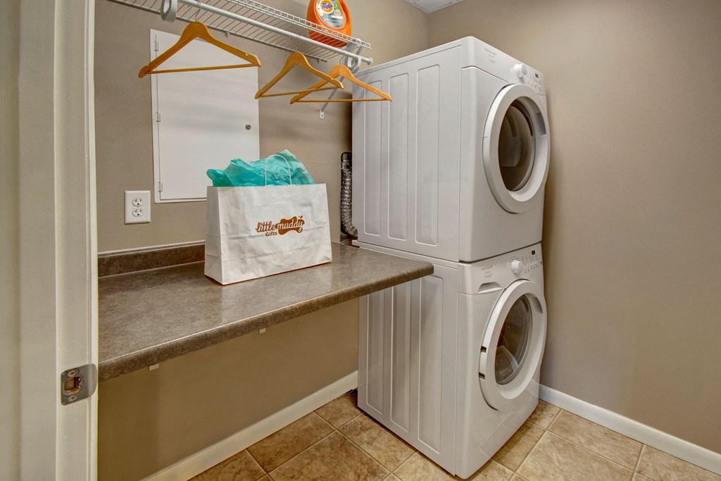 Stacked white washer and dryer in a laundry room here at Dakota Commons Apartment Homes with a tiled floor. A brown counter holds a gift bag and hangers are on shelves above, creating a tidy, organized space.