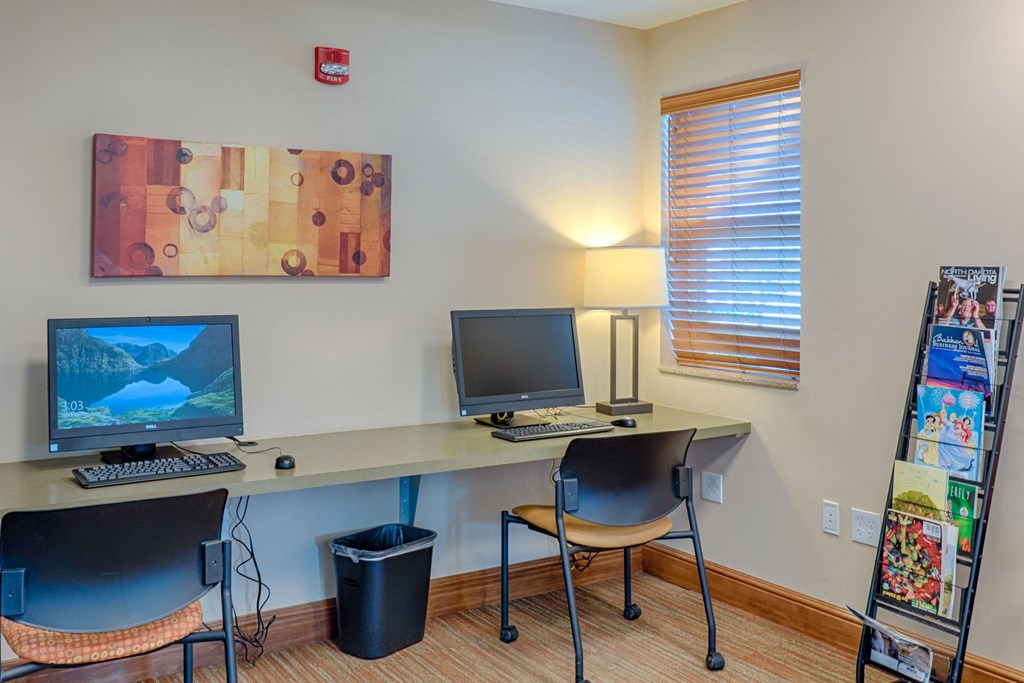 A small office space here at Dakota Ridge Apartment Homes with two computers on a desk, a lamp, and a window with blinds. A magazine rack stands to the right, while a colorful abstract painting adorns the wall, creating a calm and organized atmosphere.