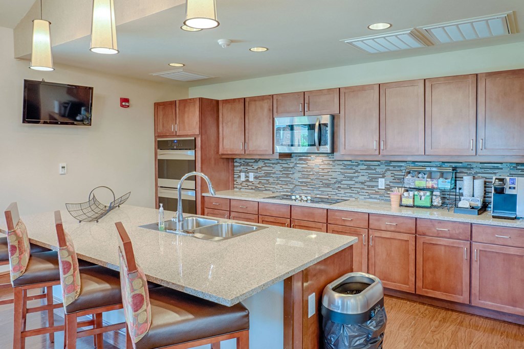 Modern kitchen here at Dakota Ridge Apartment Homes with wooden cabinets, tiled backsplash, and stainless steel appliances. Granite island counter has three chairs and a fruit basket.