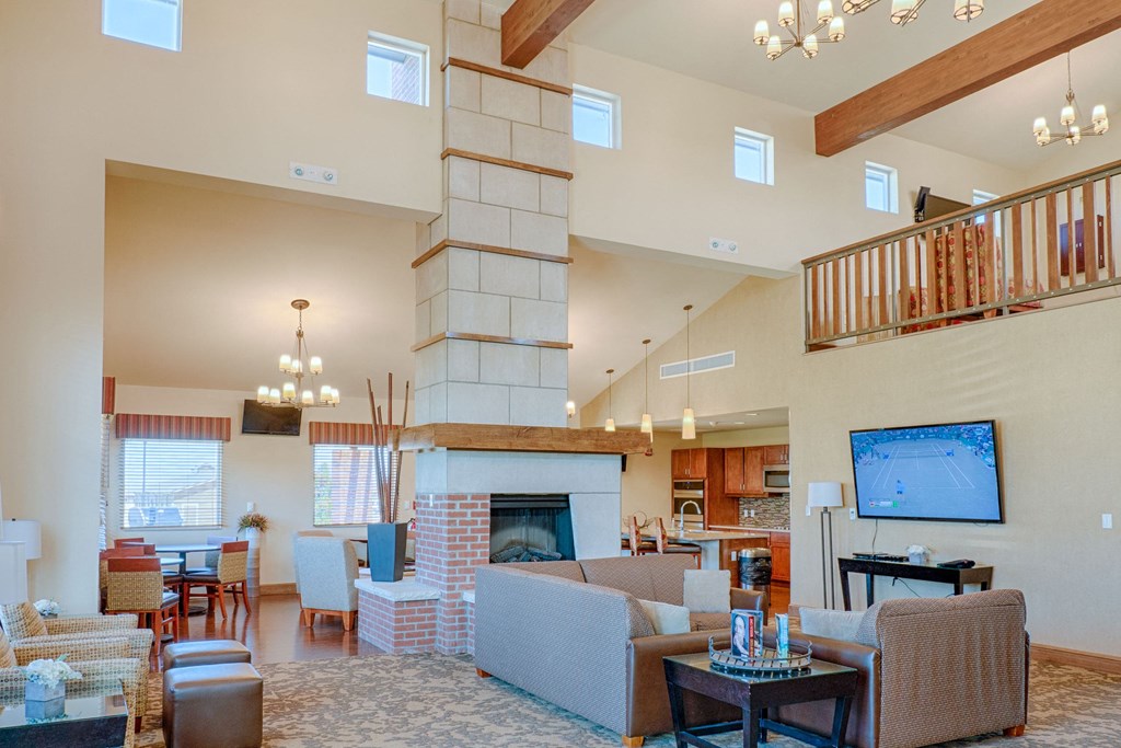Spacious living room here at Dakota Ridge Apartment Homes with high ceilings, stone fireplace, and wooden beam accents. Cozy seating area, chandelier lighting, and a TV displaying a tennis match.
