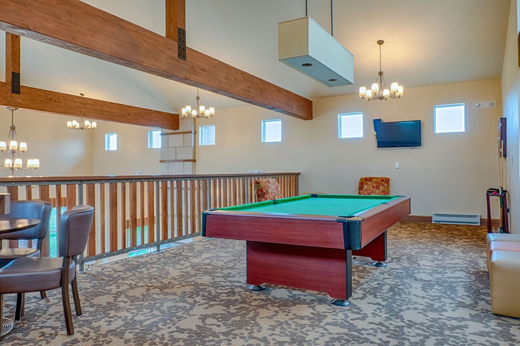 Spacious game room here at Dakota Ridge Apartment Homes with a green-felt pool table under warm lighting. Exposed wooden beams, carpeted floor, TV, and seating area create a cozy ambiance.