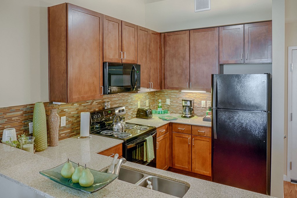 Modern kitchen here at Dakota Ridge Apartment Homes with wood cabinets, black appliances, and beige countertops. Decorative vases and artificial pears add a cozy, inviting touch.