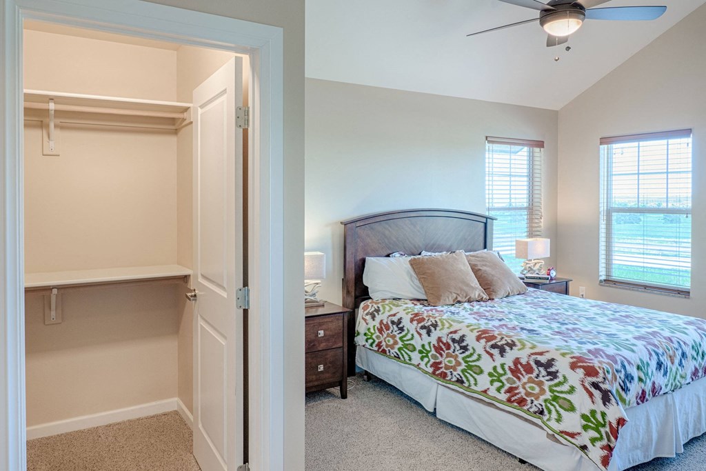 Cozy bedroom here at Dakota Ridge Apartment Homes with a colorful floral bedspread, wooden headboard, and bedside table. Open closet with shelves. Warm light streams through large windows.