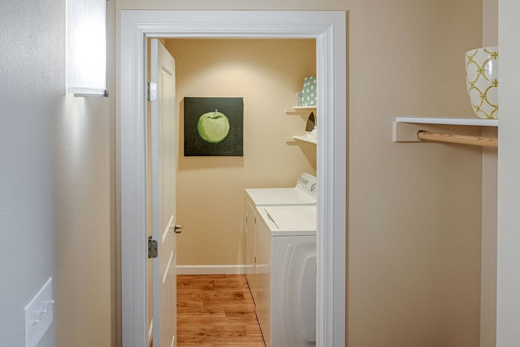 A laundry room here at Dakota Ridge Apartment Homes with beige walls features a washer and dryer. A painting of a green apple hangs above, and shelves with decorative items are visible. The mood is clean and organized.