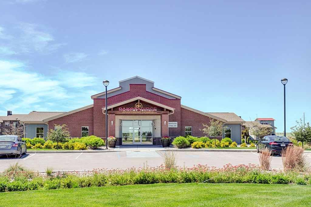 Red-brick Dakota Ridge Apartment Homes building under clear blue sky, with manicured shrubs and parked cars in front, conveying a welcoming and orderly atmosphere.