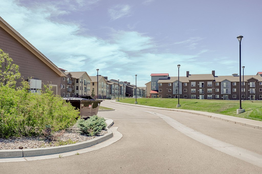 Wide suburban street with modern Dakota Ridge Apartment Homes buildings on the right. Lush greenery and a clear blue sky create a calm, spacious suburban feel.