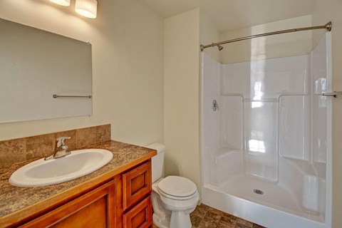 Compact bathroom here at Phoenix Ridge Apartment Homes with a white shower, beige walls, and tiled floor. Features a wooden vanity, brown countertop, sink, and toilet, under warm lighting.