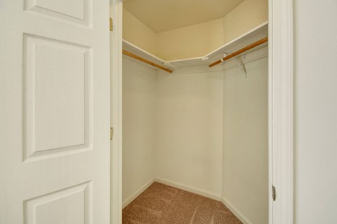 Empty walk-in closet here at Phoenix Ridge Apartment Homes with beige walls, carpeted floor, and wooden hanging rods on white shelves. The open door reveals a spacious, clean feel.