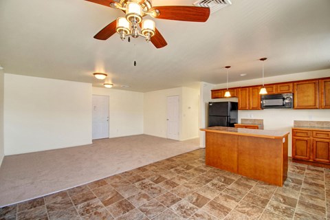 Open-concept room here at Phoenix Ridge Apartment Homes with beige walls and dual flooring: carpeted area leads to a tiled kitchen with wooden cabinets, an island, and a ceiling fan. Cozy and spacious.