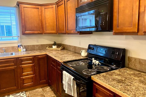 A cozy kitchen here at Phoenix Ridge Apartment Homes with warm wooden cabinets, granite countertops, a black stove with a striped towel, and a plant by a bright window on a tiled floor.